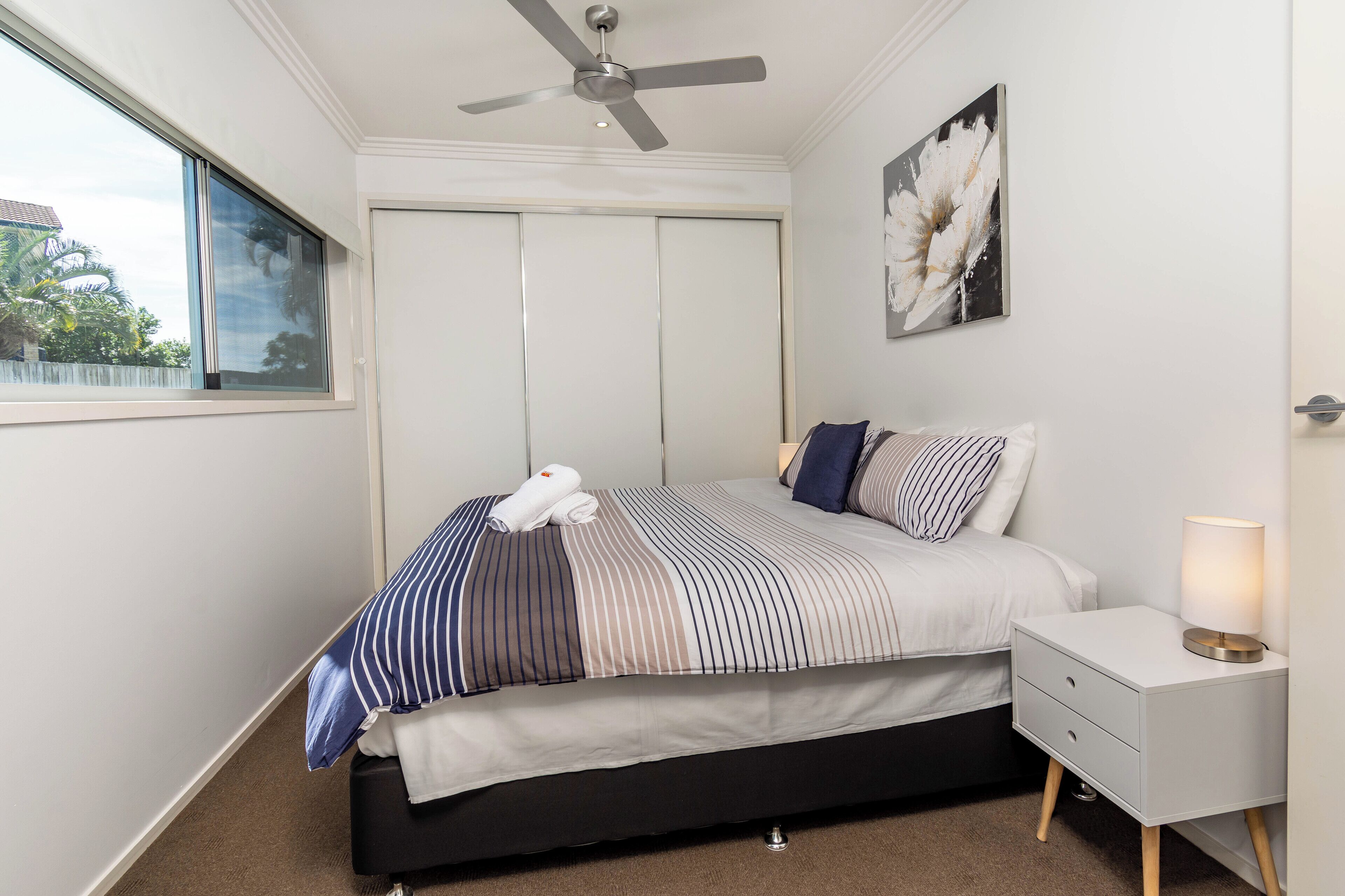 A bedroom with a bed, a nightstand, a ceiling fan, a window, and a wall-mounted artwork.