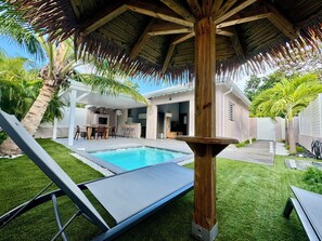 Un espace aménagé au bord de la piscine, comprenant une pergola en bois, un fauteuil de relaxation et un coin repas avec une table et des chaises.