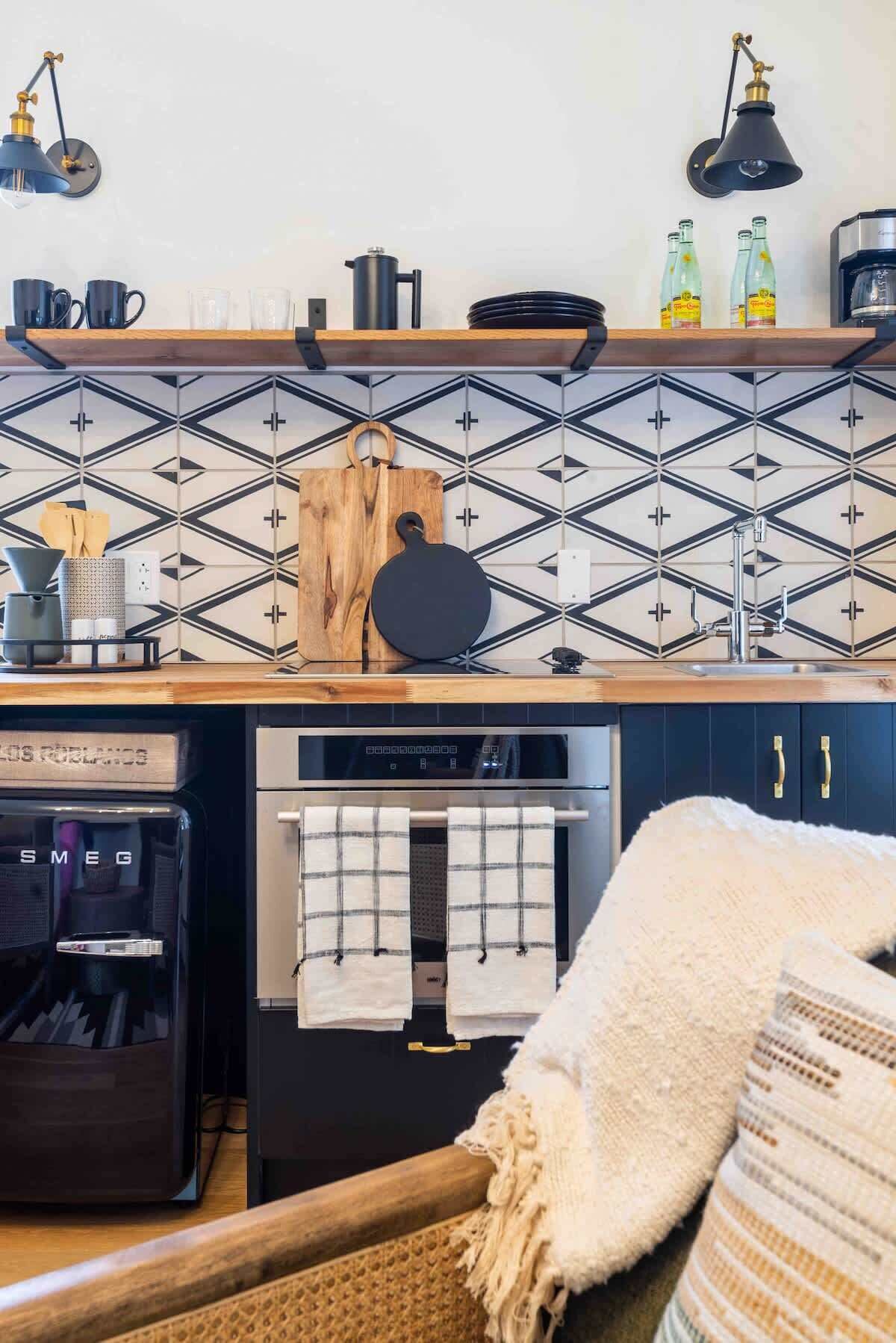 A kitchen with a black and white patterned backsplash, a wooden cutting board, a kettle, and a coffee machine.