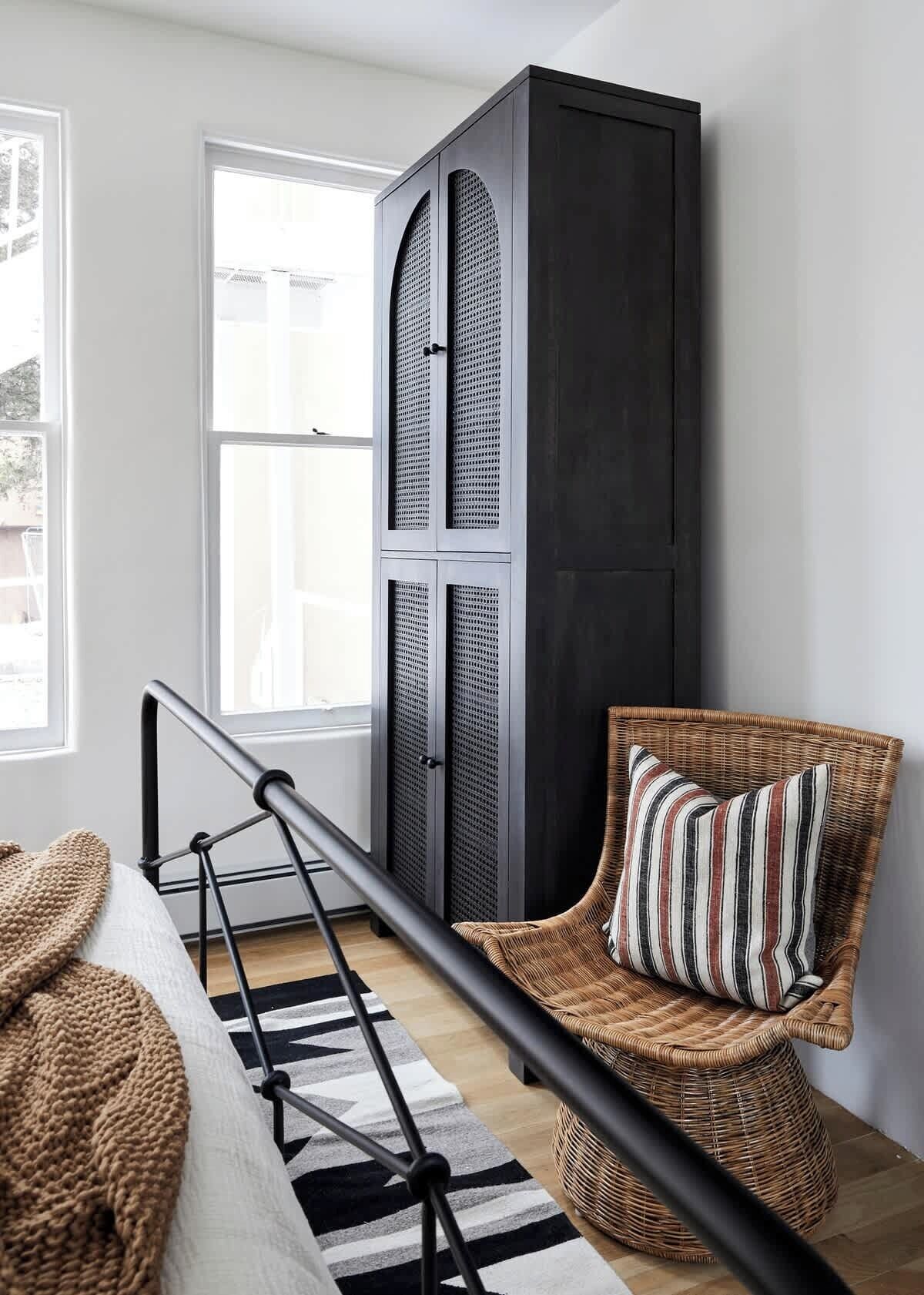 Ein Zimmer mit einer schwarzen Metalltreppe, einem Korbstuhl mit gestreiftem Kissen und einem großen, dunklen Schrank an einer weißen Wand.