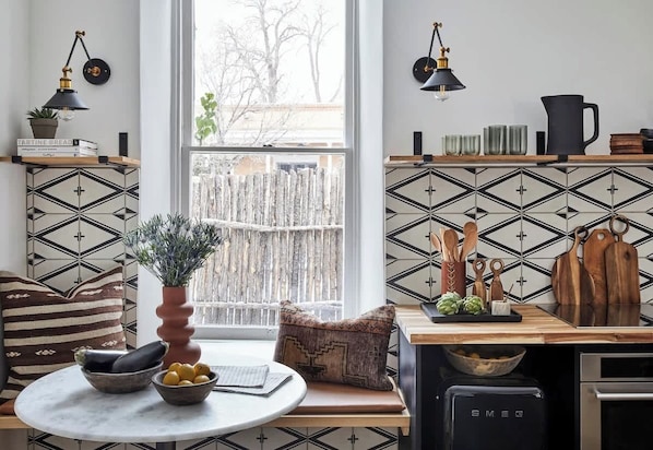A kitchen with a black and white geometric backsplash, a wooden table with a bowl of fruit, a vase with greenery, and a black refrigerator.