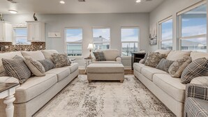 A spacious living room with a beige sofa set, a matching ottoman, and a large window providing a view of the city. - Amazing Outdoor Space, Beautiful Interior with King Beds, Easy Walk to Beach! (Crystal Beach)