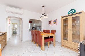 Uma área de refeições com mesa de madeira e cadeiras vermelhas, um armário com portas de vidro e uma cozinha visível por uma abertura.
