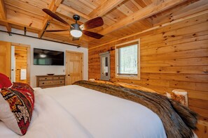 Une chambre dans un chalet en bois, avec un lit, un ventilateur de plafond, une télévision et une fenêtre avec des stores.