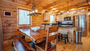 Ein Holzblockhaus-Interieur mit einem Holzesstisch, Stühlen, einem Barhocker, einer Pendelleuchte, einer Mikrowelle, einem Kühlschrank und einem Mülleimer.