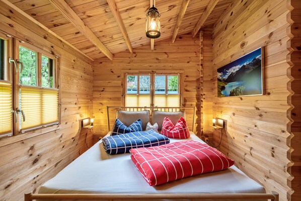 A wooden cabin bedroom with a bed, two windows, a wall-mounted picture, and a ceiling light.