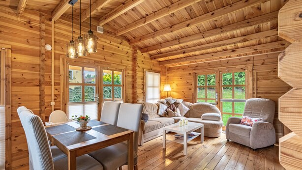Ein Holzhäuschen-Interieur mit Esstisch, Sofa, Sessel, großen Fenstern und Holzbalkendecke.