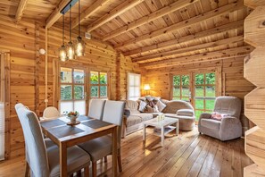 A wooden cabin interior with a dining table, sofa, and armchair, large windows, and a wooden ceiling.