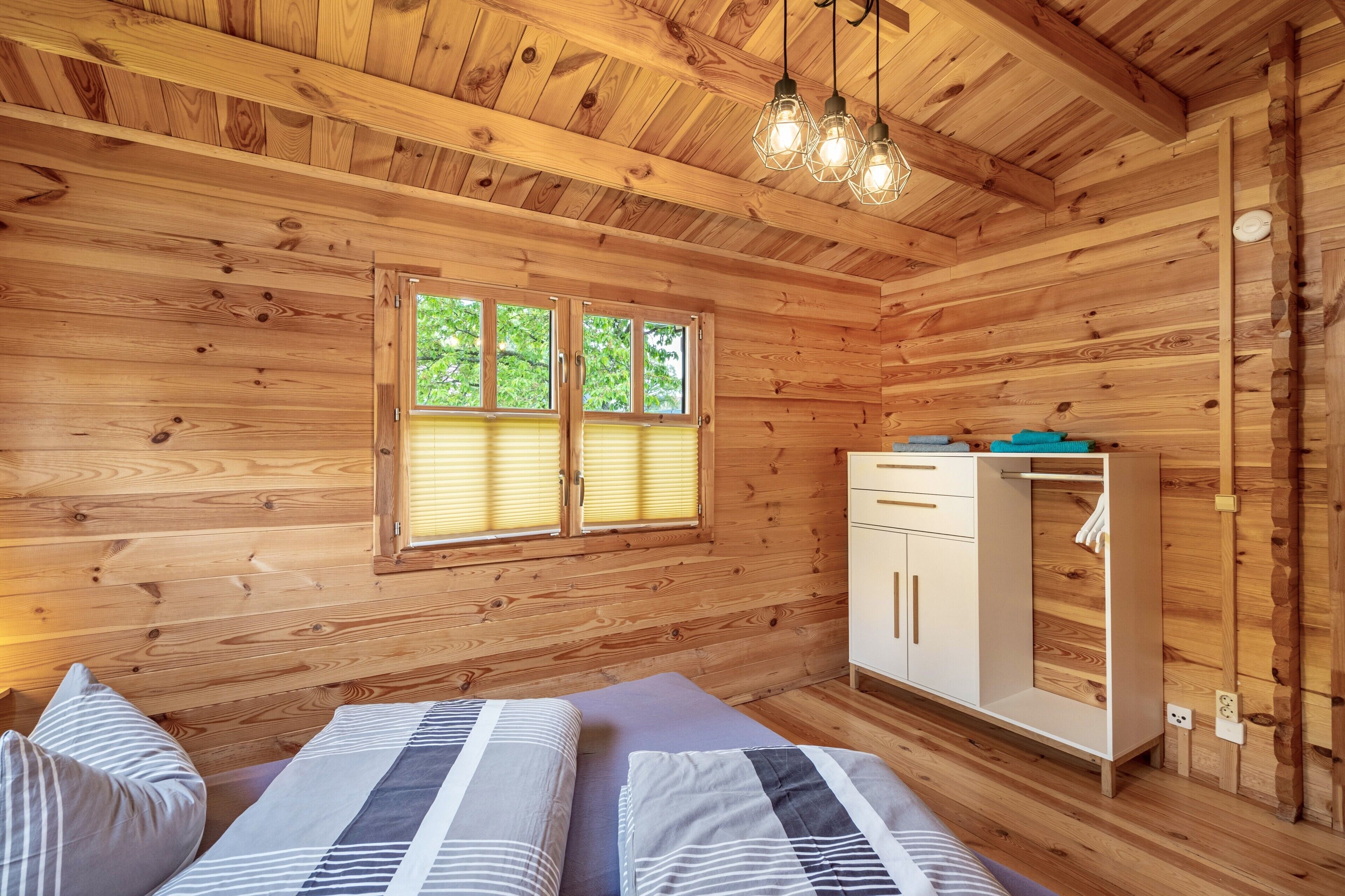 A wooden cabin room with two beds, a white wardrobe, and a window with blinds.