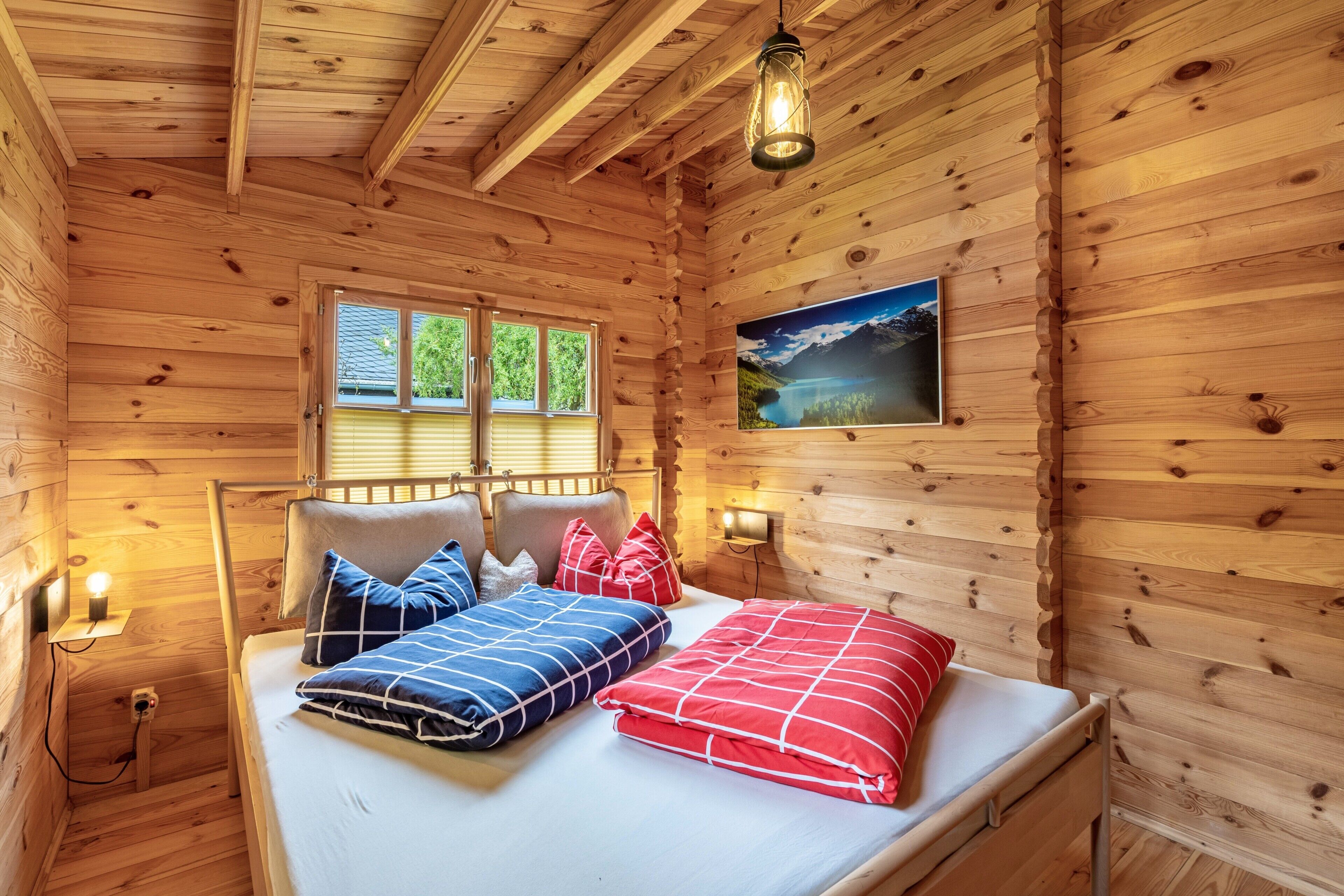 A wooden cabin room with a bed, a window, a wall-mounted picture, and a hanging light fixture.