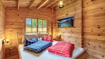 A wooden cabin room with a bed, a window, a wall-mounted picture, and a hanging light fixture.