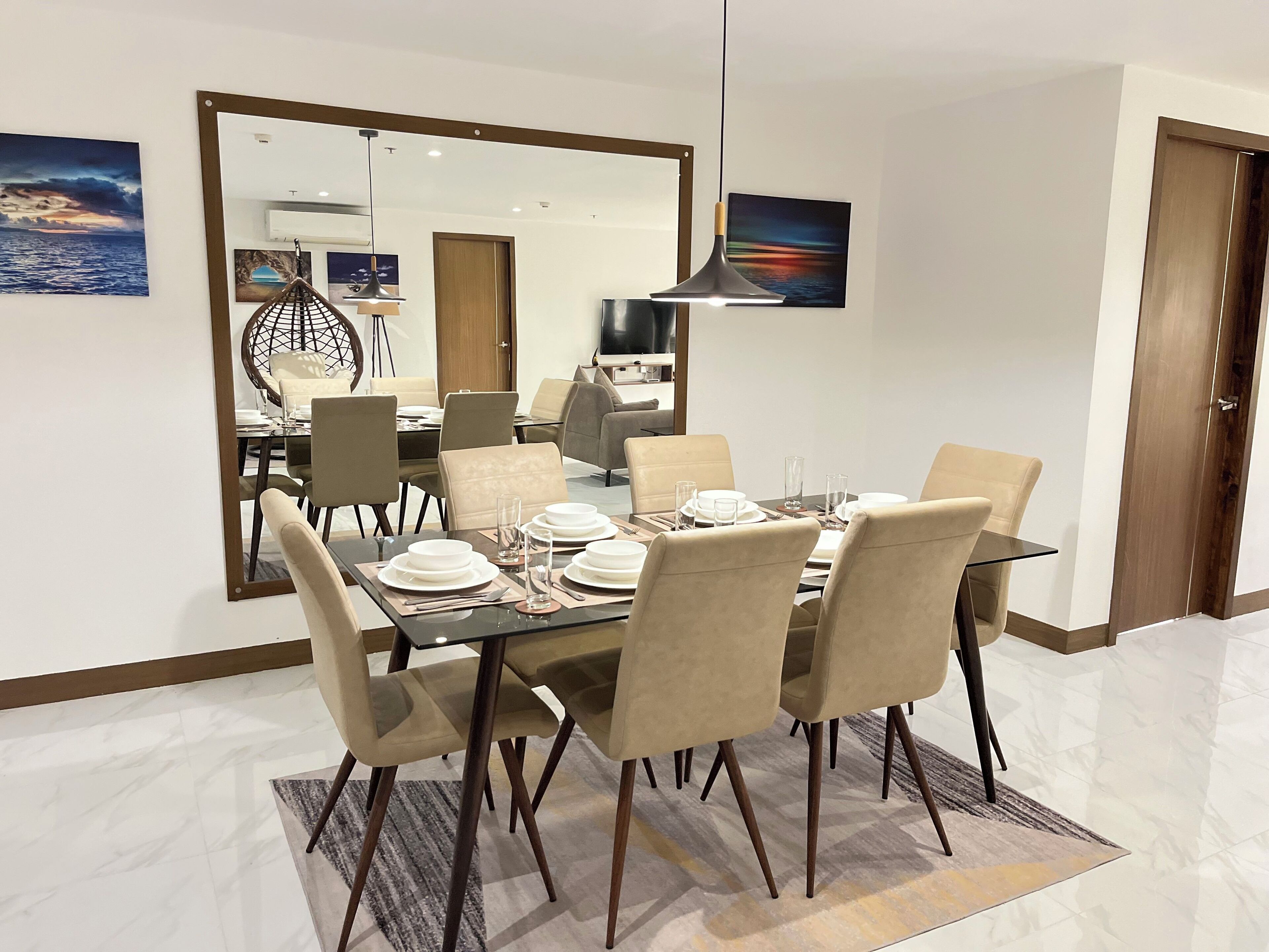 A dining area with a rectangular table set for six, surrounded by six chairs. There is a large mirror on the wall, a hanging pendant light, and a television in the background.