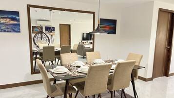 A dining area with a rectangular table set for six, surrounded by six chairs. There is a large mirror on the wall, a hanging pendant light, and a television in the background.