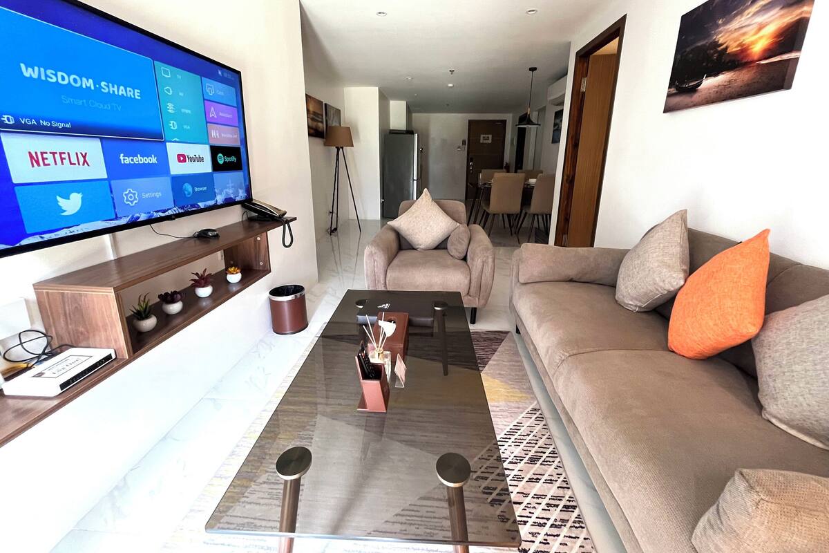 Een moderne woonkamer met een flatscreen televisie waarop een slimme tv-interface wordt weergegeven, een bank met kussens, een salontafel, en een eethoek op de achtergrond.