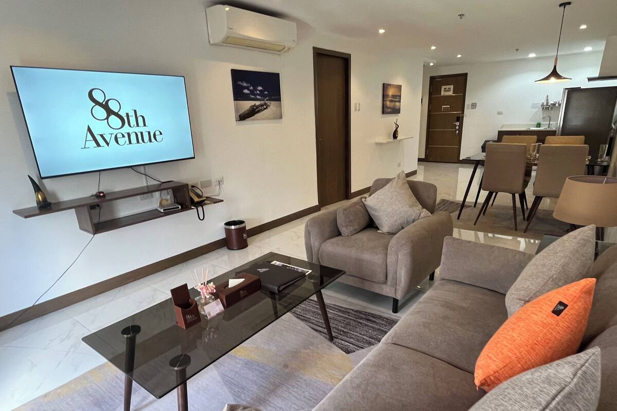 Een moderne woonkamer met een flatscreen televisie waarop “8th Avenue” staat, een bank, een salontafel en een eethoek op de achtergrond.