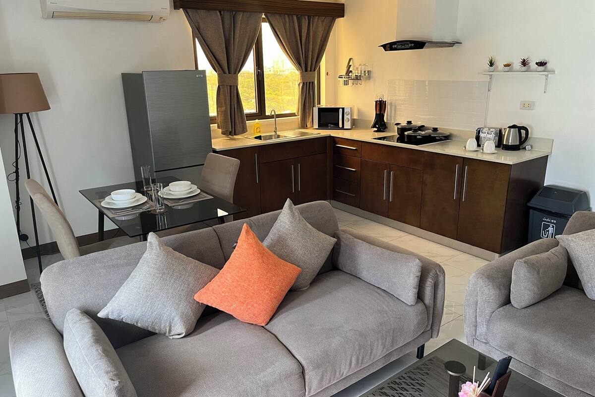 Een moderne woonkamer met een grijze bank, een eettafel met stoelen, een keuken met een magnetron en een raam met gordijnen.
