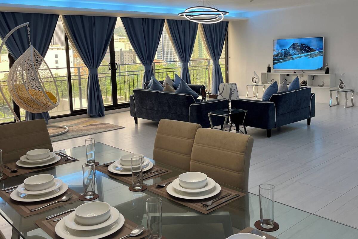 Een moderne eetruimte met een glazen tafel voor vier personen, een bank en een aan de muur bevestigde televisie.
