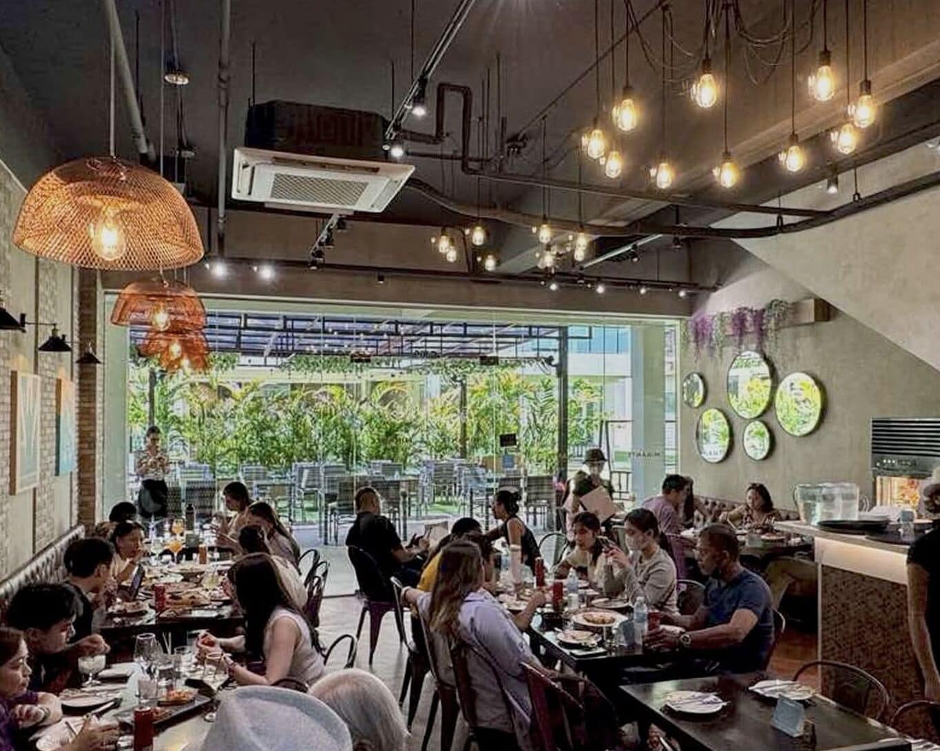 Een restaurant met meerdere eettafels, hangende verlichtingsarmaturen en uitzicht op groen buiten.