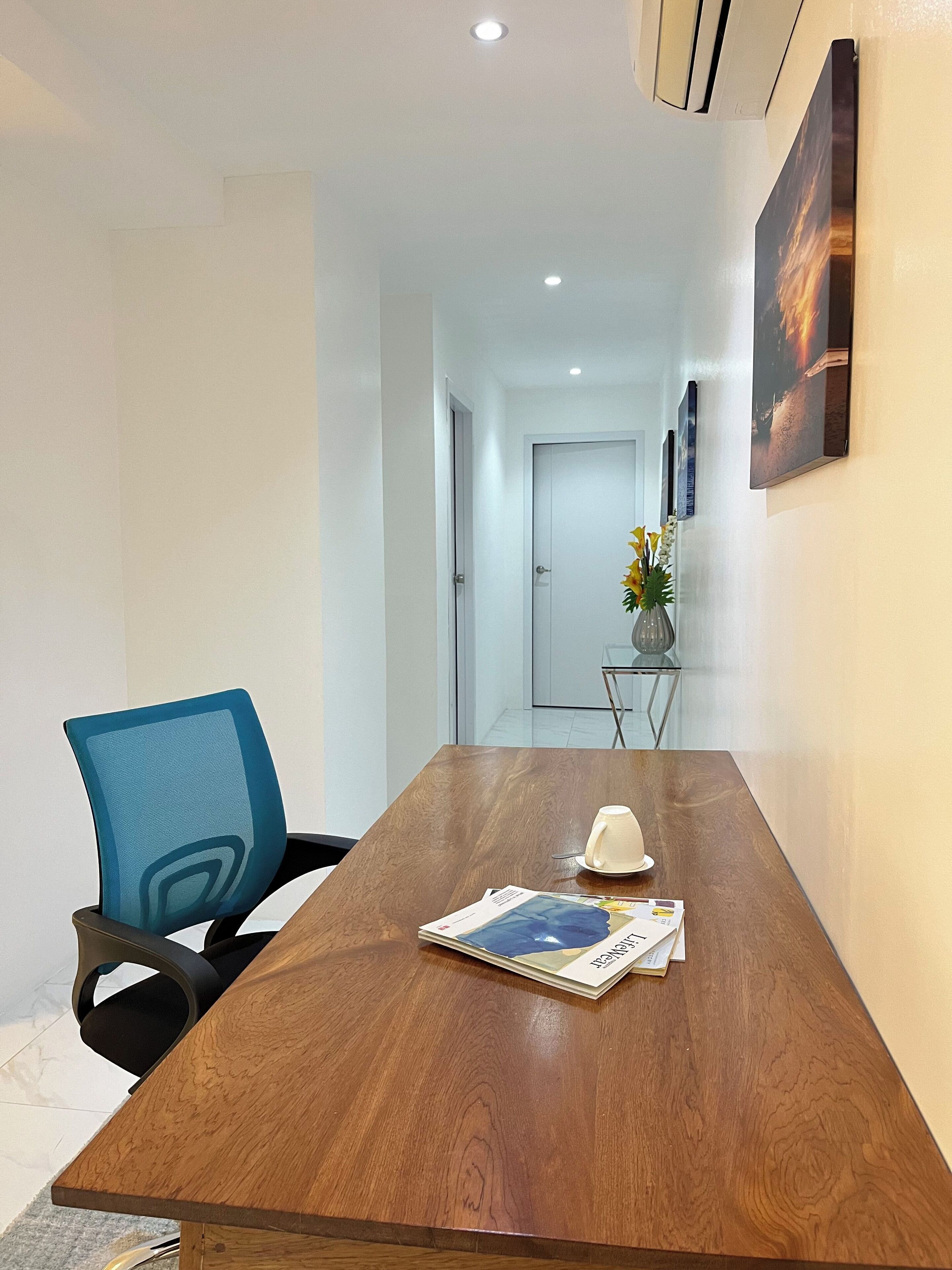 A conference room with a wooden table, a blue office chair, a white vase with flowers, and a framed picture on the wall.