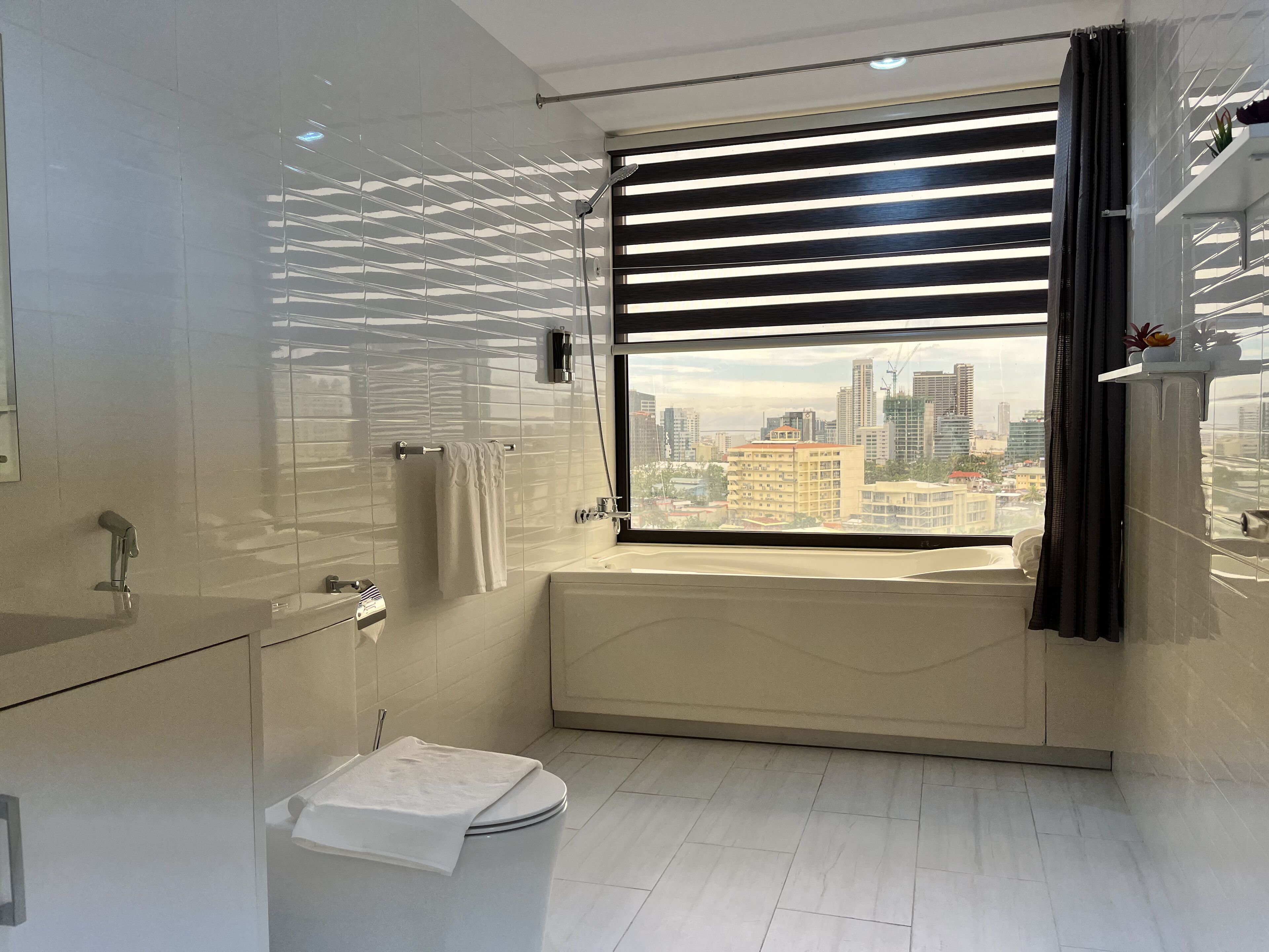 Un bagno moderno con una vasca, un water e un lavandino. C'è una finestra con persiane e vista sulla città.