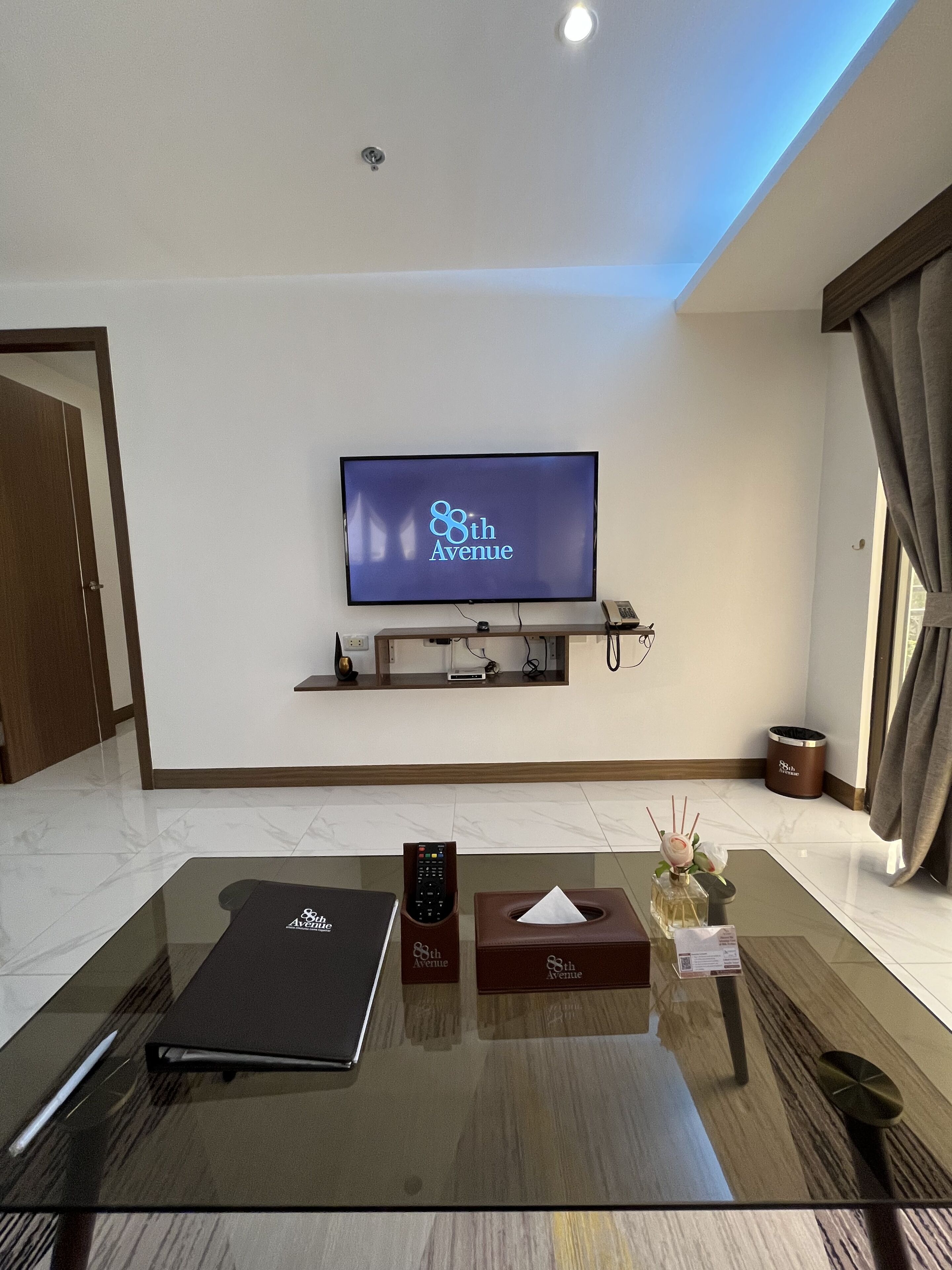 A modern living room with a flat-screen TV mounted on the wall, a glass coffee table, a laptop, a remote control, a tissue box, and a decorative vase.