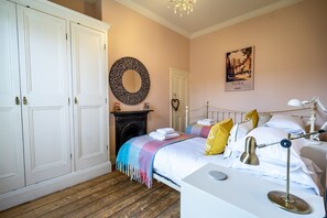 Une chambre à coucher comprenant un lit, une table de chevet, une lampe, une armoire, une cheminée et un mur orné d’un miroir rond et d’une affiche.