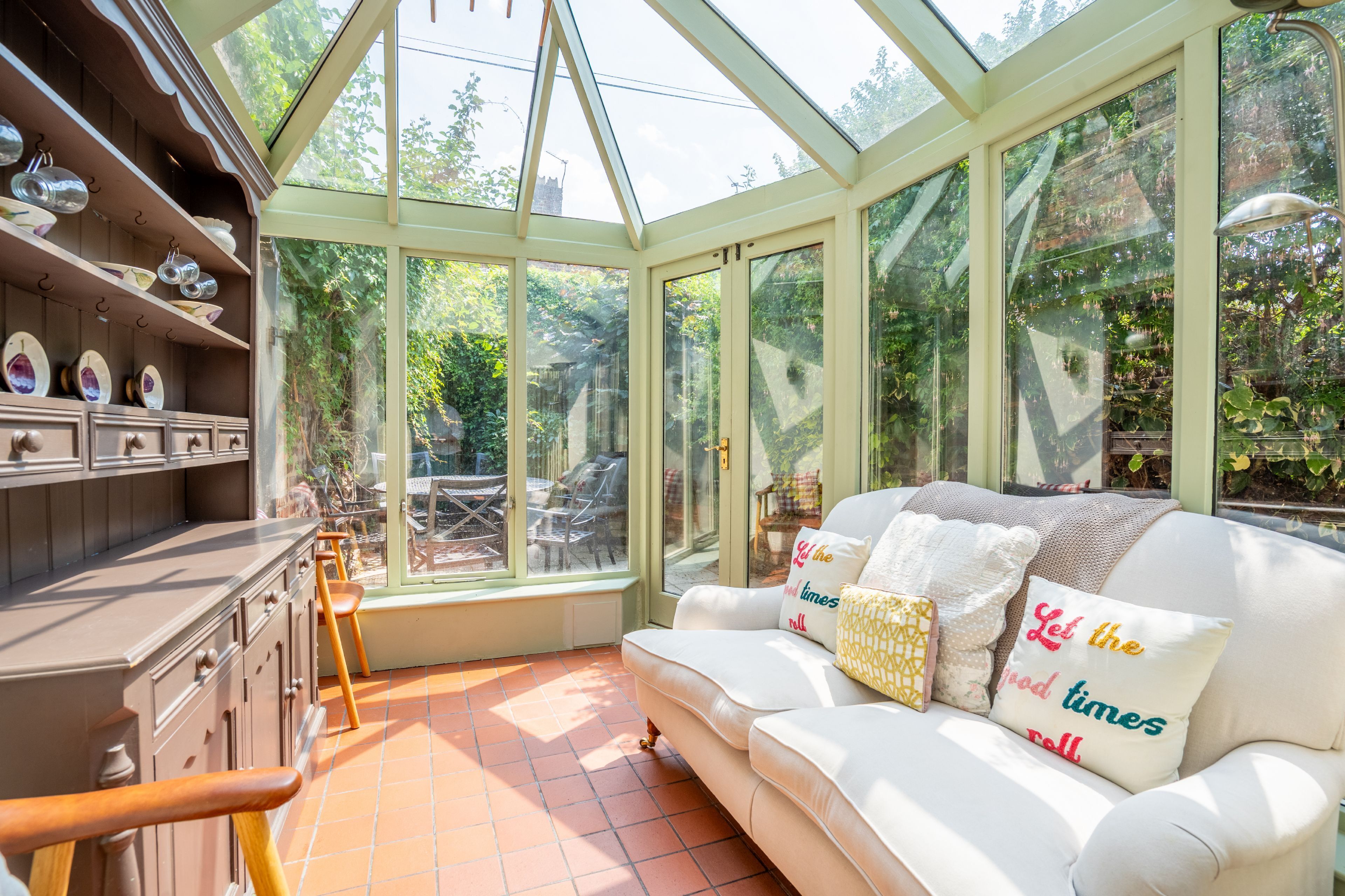 A conservatory with a glass ceiling, a white sofa with decorative pillows, and a wooden cabinet with dishes.