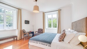 Un dormitorio con una cama grande, un escritorio, dos sillas, un banco y un televisor en la pared.