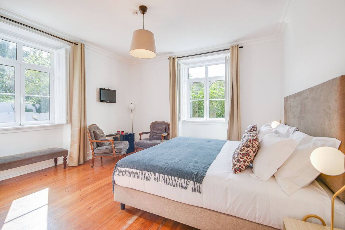 Quarto com uma cama grande, uma escrivaninha, duas cadeiras, um banco e uma televisão na parede.