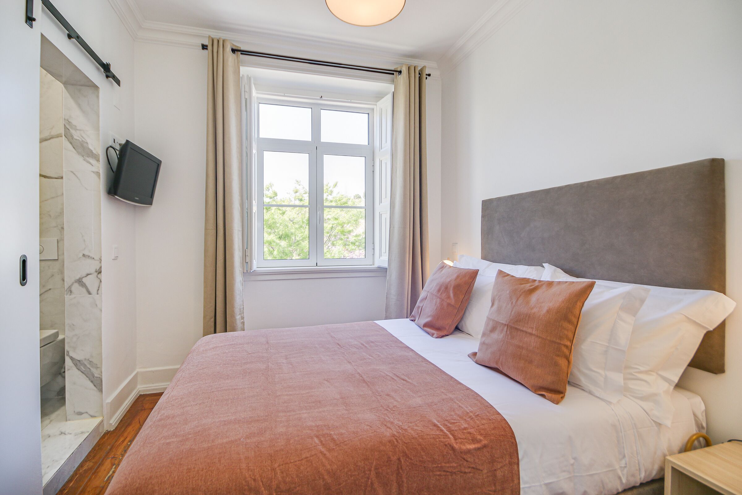 Ein Schlafzimmer mit einem großen Bett, einem an der Wand montierten Fernseher, einem Fenster mit Vorhängen und einem Duschbereich aus Marmor.