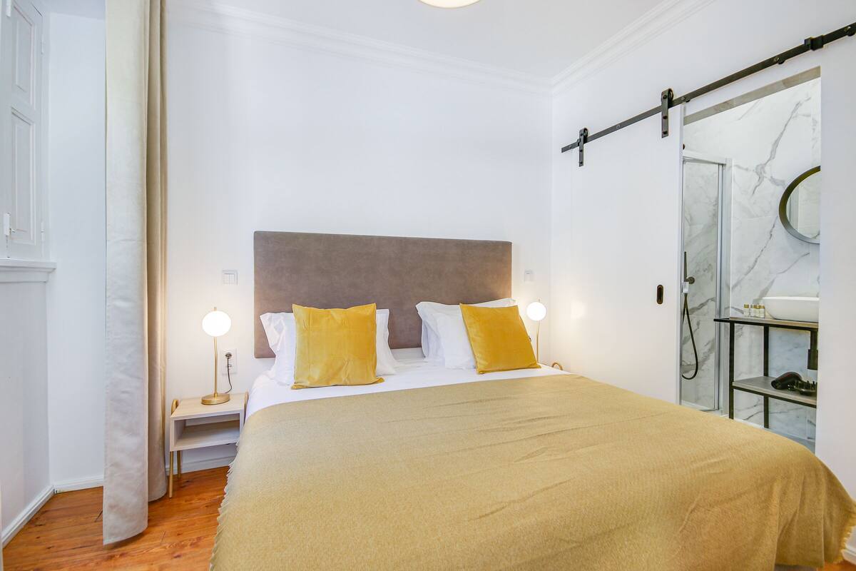 Quarto de hotel bem arrumado, com uma cama grande, duas luminárias de cabeceira, encosto para a cama e banheiro com parede de mármore e pia.