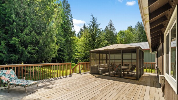 A wooden deck with a glass enclosure, outdoor furniture, and a view of a forested area. - Mt. Rainier Cabin-Hot Tub-AC-Quiet (Packwood)