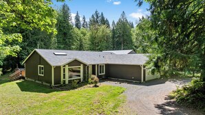 A two-story house with a covered porch and a garage, surrounded by dense forest. - Mt. Rainier Cabin-Hot Tub-AC-Quiet (Packwood)