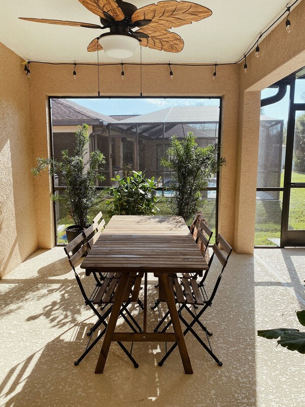 Eine sonnendurchflutete Terrasse mit Holztisch und -stühlen, einem Deckenventilator und Blick auf ein Haus und grüne Pflanzen.