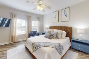 A bedroom with a large bed, a ceiling fan, two wall-mounted paintings, a nightstand, and a TV on the wall. - Top Floor/Corner Unit - Waterfront, Pool, Hot Tub, Steps to the Beach, + Tickets to Fun Activities! (Treasure Island)