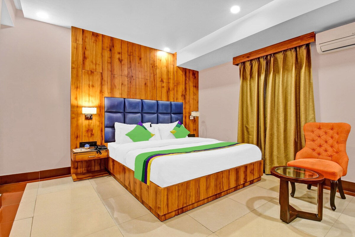 A hotel room with a wooden headboard, a bed with white bedding and colorful pillows, a nightstand with a lamp, a glass table with an orange chair, and a gold curtain.
