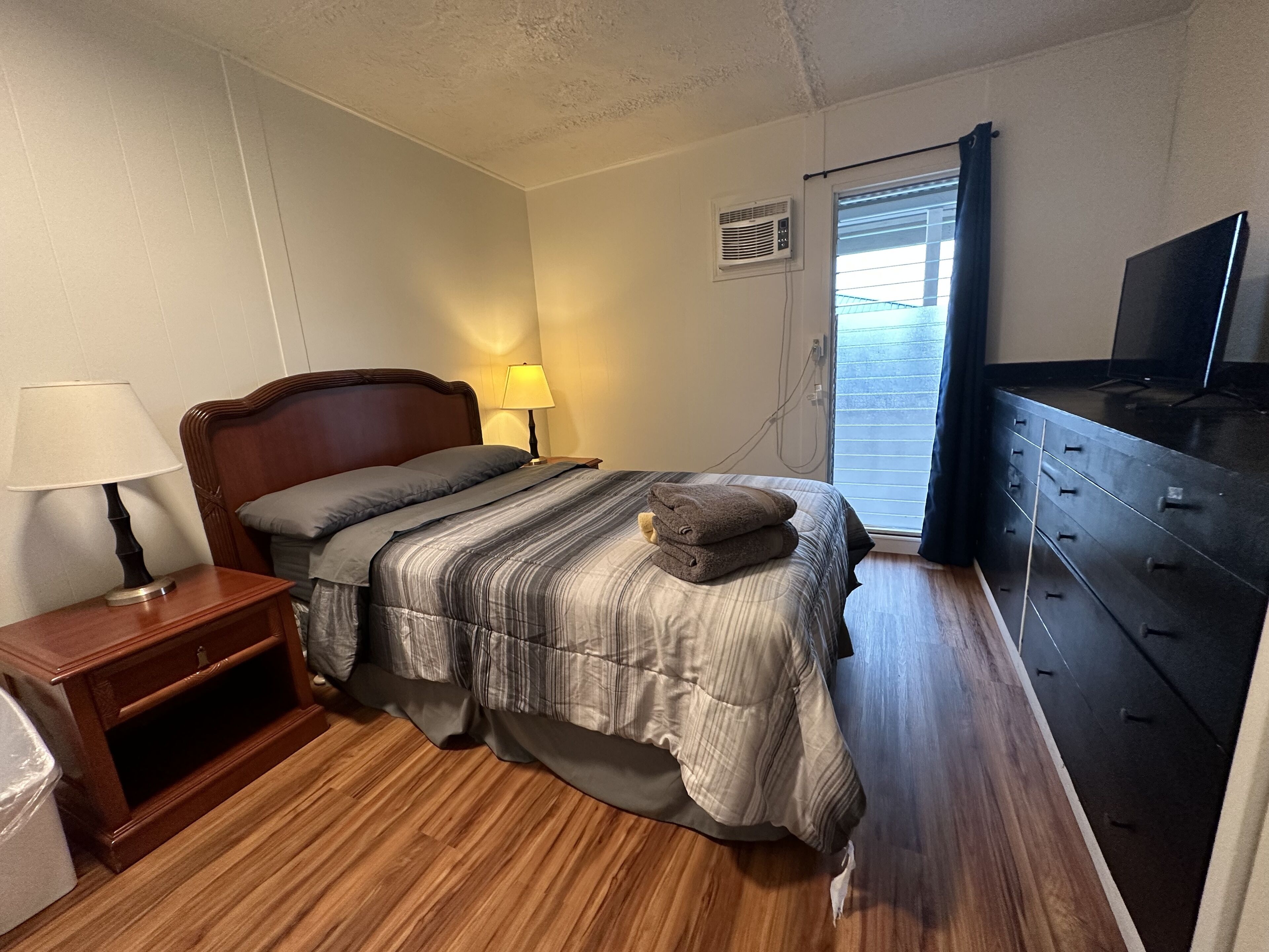A bedroom with a wooden bed, a nightstand with a lamp, a dresser, and a television on a stand.
