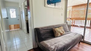 A room with a grey sofa, a wooden floor, a white door, and a view of the outside through a glass door. -  Unique getaway, 3-bedroom tropical paradise in the heart of Hilo town (Hilo)