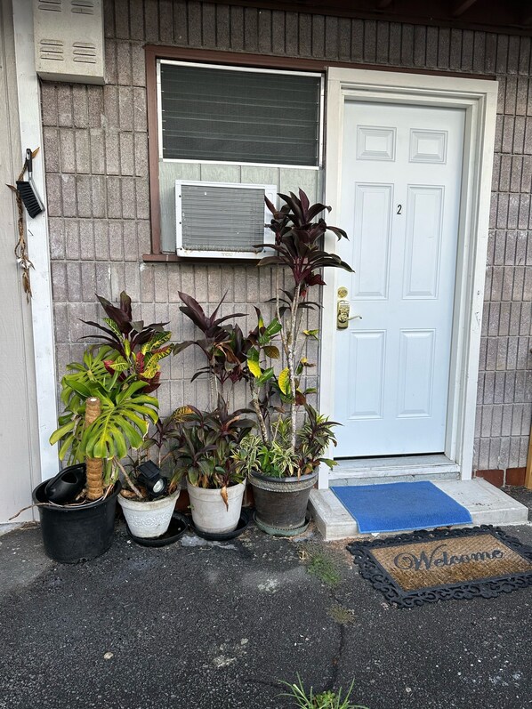 A house entrance with a white door, a window with a closed shutter, and several potted plants in front. - Rainbow Hale, Unit 2—a delightful and convenient pet-friendly retreat. (Hilo)