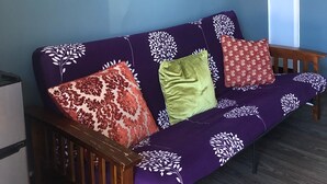 Ein Zimmer mit einem violetten Sofa, verziert mit weißen floralen Mustern, einer Holzbank und einem Wandbild, das eine Welle zeigt.