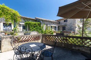 Un bâtiment en pierre doté d’un patio comprenant une table ronde et des chaises, un parasol et des plantes en pot.