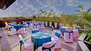 Un área de comedor al aire libre con mesas y sillas redondas, cubierta con manteles azules y sillas blancas adornadas con cintas azules.