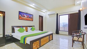 A hotel room with a bed, a nightstand, a chair, a painting, and a window with a view. - Treebo Amaya Inn Sector 72 (Noida)