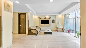 A modern hotel room with a sofa, a small table, a TV mounted on the wall, a Buddha statue, and a large window with plants. - Treebo Sai Village Gold Souk - Sec 43 (Gurugram)