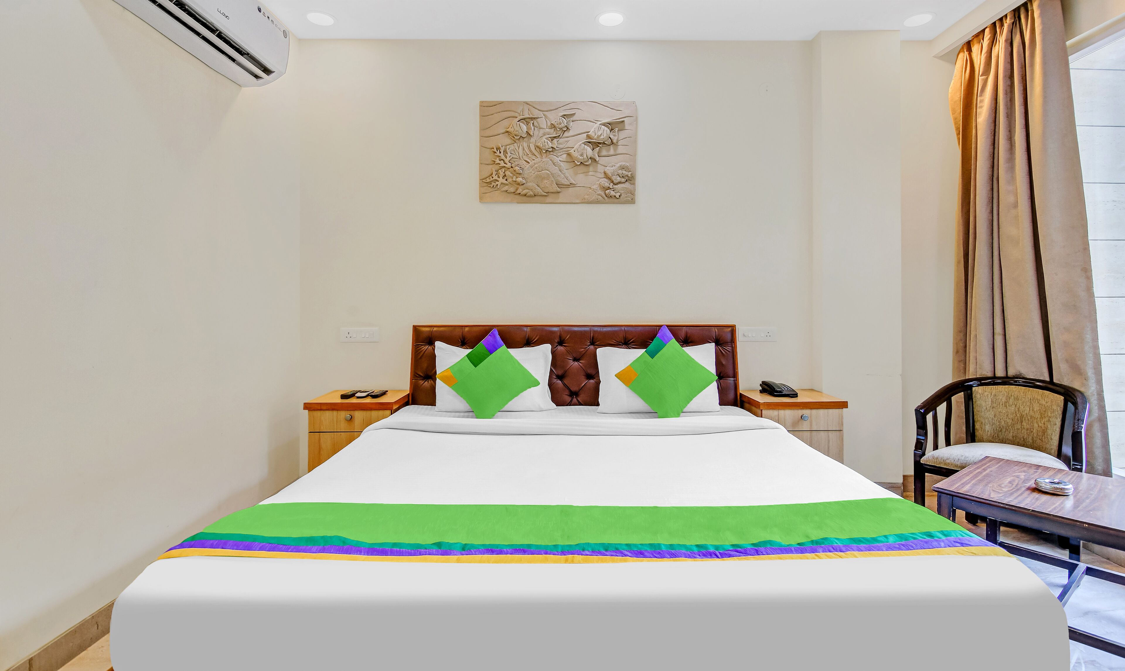 A hotel room with a bed, a nightstand, a chair, a table, an air conditioning unit, and a painting on the wall.