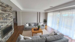 Ein Wohnzimmer mit Kamin aus Stein, zwei grauen Sofas, einem Holztisch und einem großen Fenster mit transparenten Vorhängen.