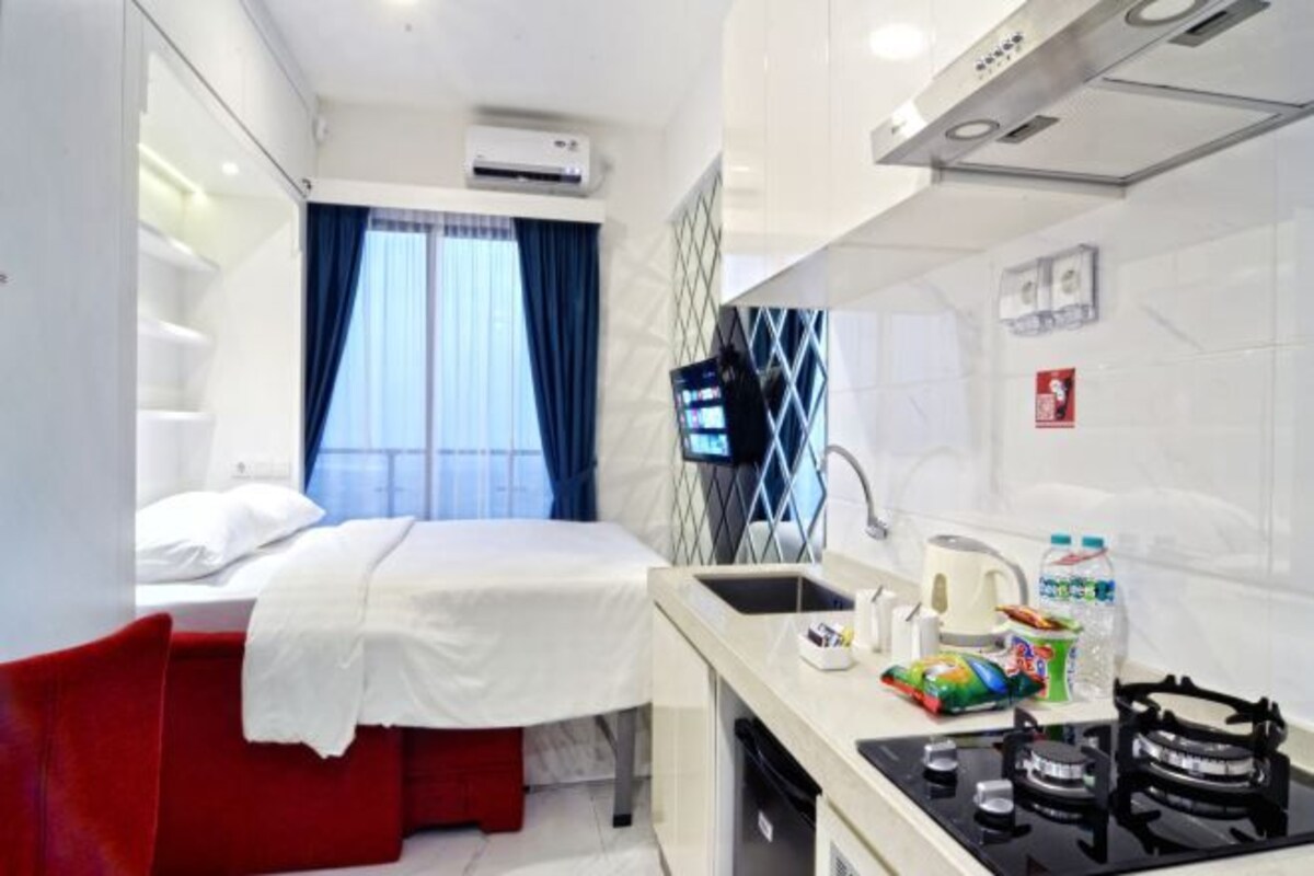 A compact hotel room with a bed, a small kitchenette, a sink, and a window with curtains.