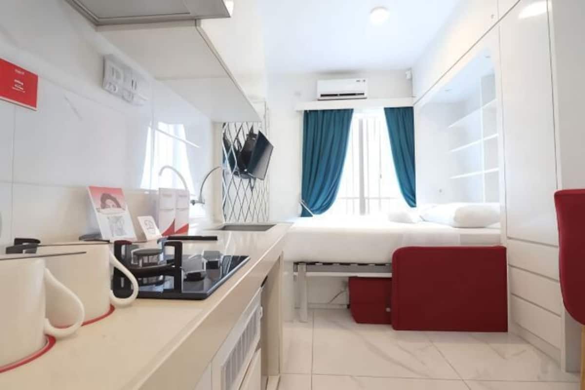 A compact hotel room with a bed, a small desk with a computer, a red chair, and a window with blue curtains.