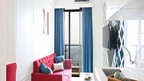 A modern hotel room with a red sofa, a small table with a cup and saucer, a wooden tray with plants, a large window with blue curtains, and a TV mounted on the wall.