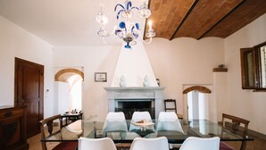 Une salle à manger avec une table en verre, des chaises blanches, un lustre bleu et blanc, une cheminée et des poutres en bois au plafond.
